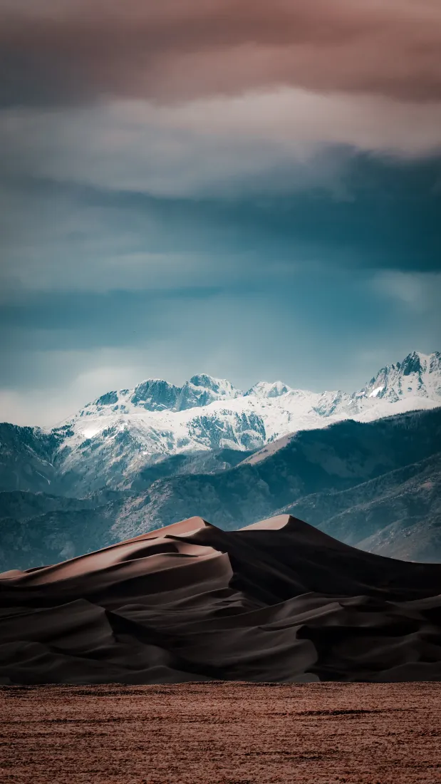 前景荒漠沙丘，中景层叠青蓝山脉，远景覆雪高峰与渐变云层。