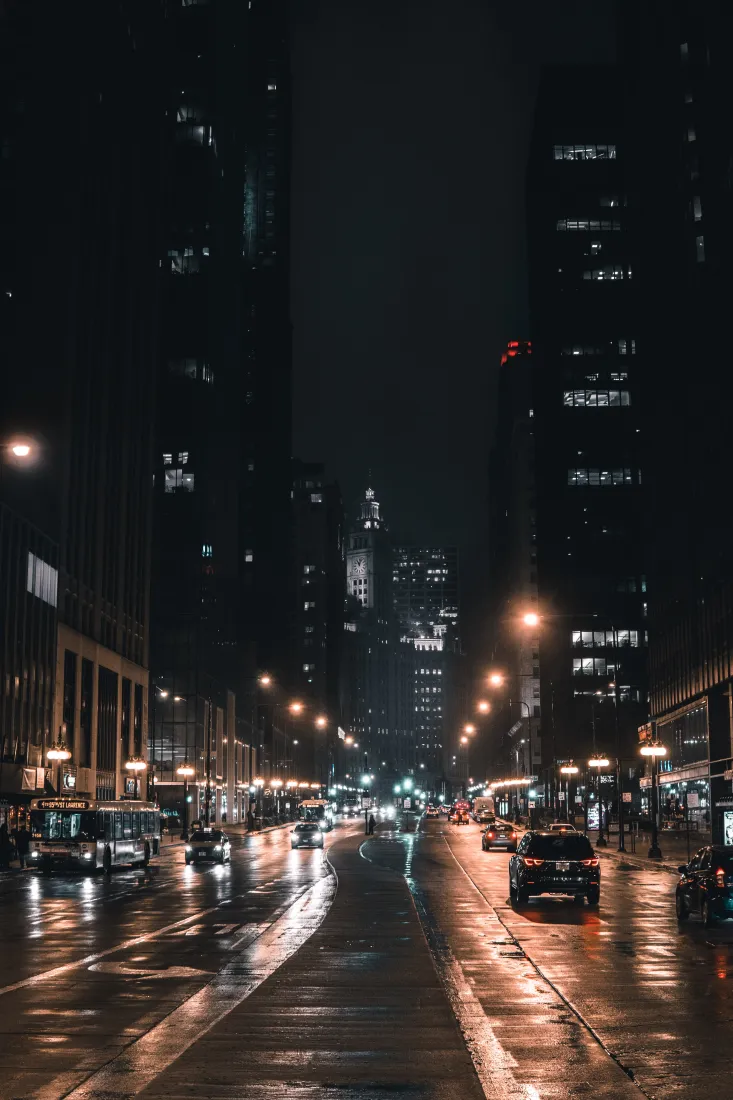雨夜城市街道，湿滑路面反射暖黄路灯，两侧高楼林立，车流穿行。
