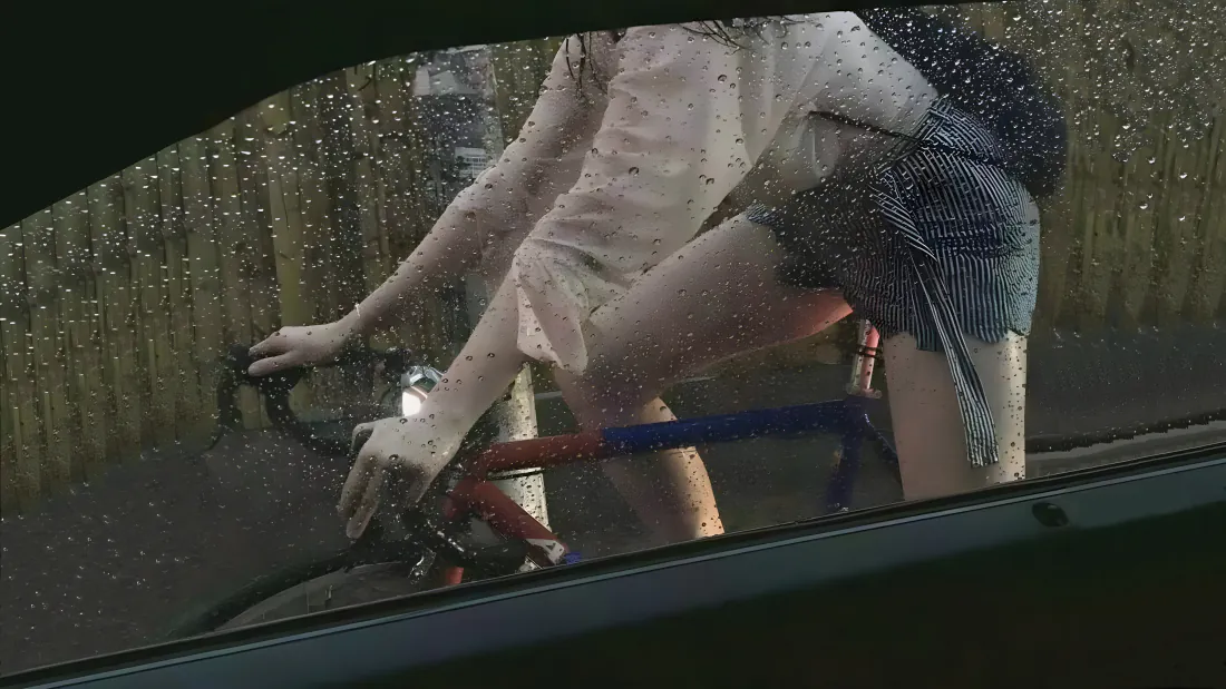 透过布满雨滴的车窗，看到一名女孩在雨天街道上骑着自行车。