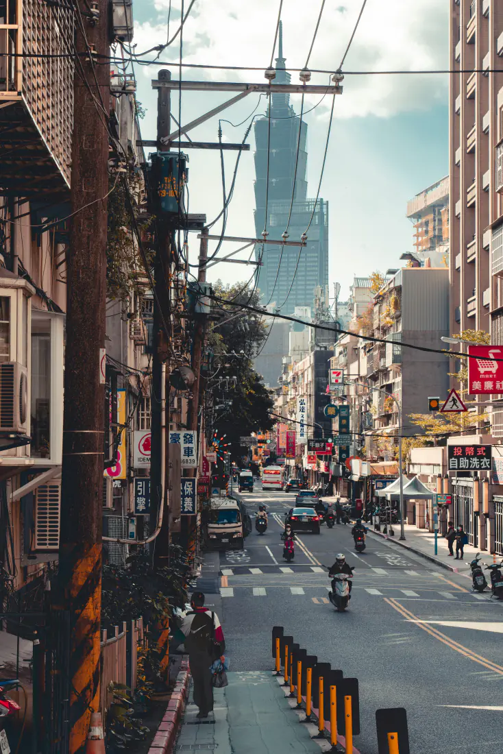 台北老城街巷向远处延伸，两侧临街建筑林立，尽头矗立台北101大楼，街道有摩托与行人。