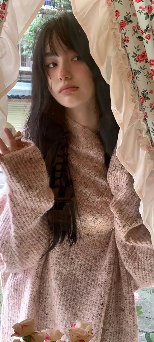 黑长直齐刘海少女身着粉色毛衣，站在碎花窗帘旁的窗边。