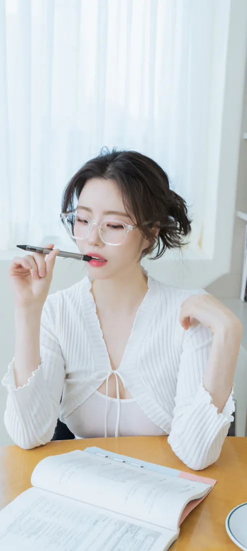 戴眼镜的白衣女生坐在书桌前，手持笔，面前摊开笔记本，背景是白色窗帘。