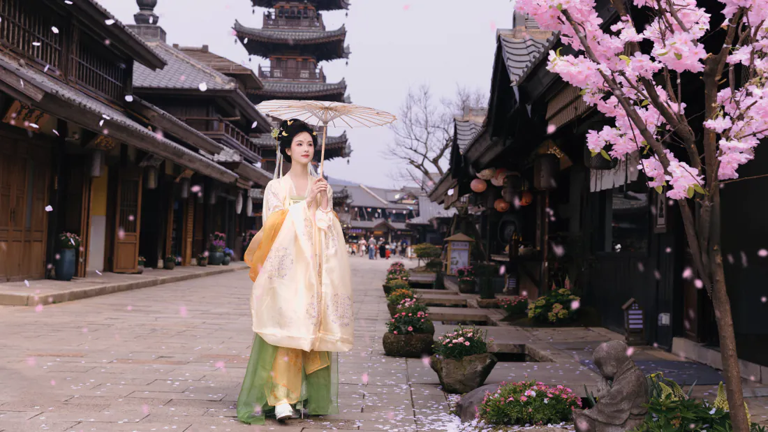 5k陈都灵壁纸|古风汉服古街背景图片 - 唯美浪漫「哲风壁纸」 陈都灵身着浅黄汉服手持油纸伞,站在飘满花瓣的古街,旁有樱花树与古塔。