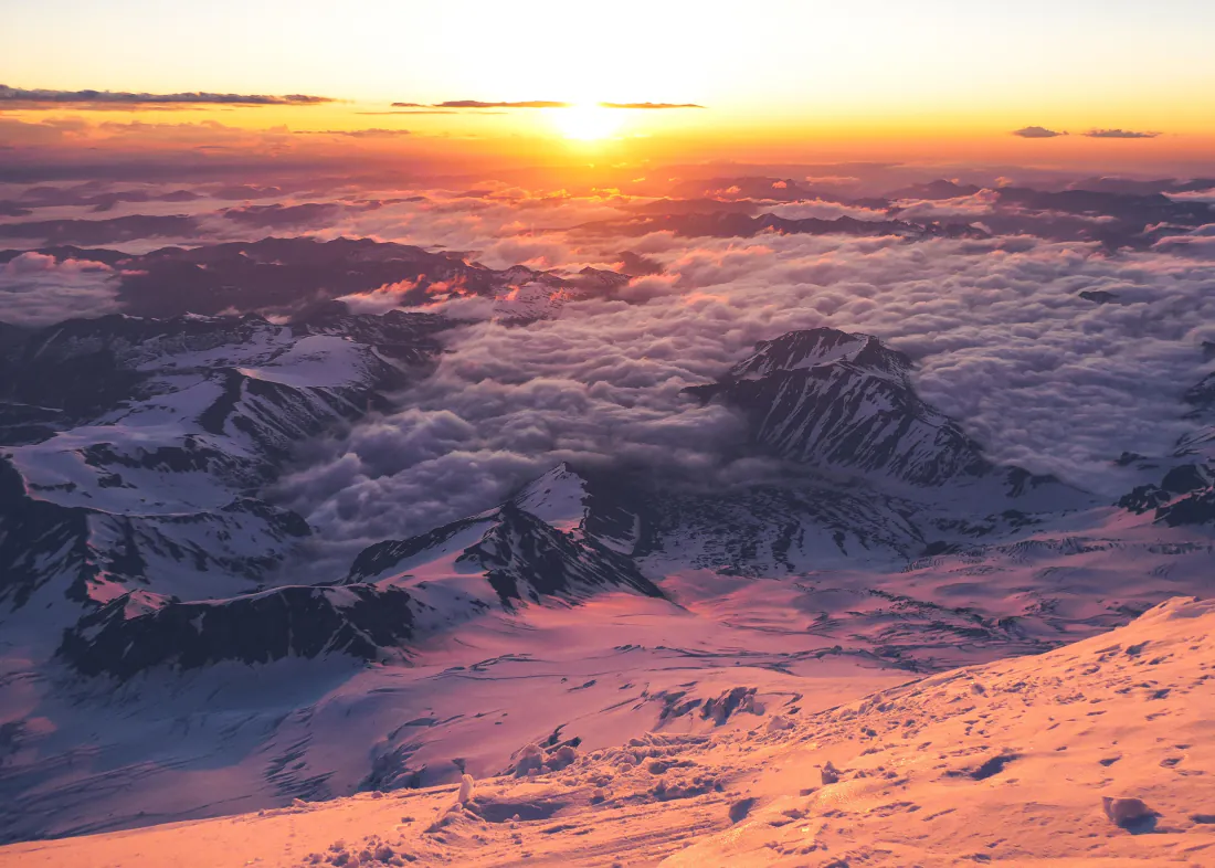 高空俯瞰日出时分的雪山群峰，云海翻涌，暖金色霞光铺满积雪。
