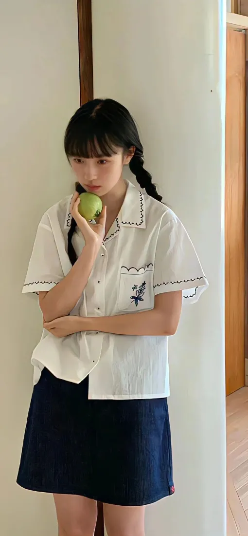 扎双麻花辫的年轻女生穿白衬衫深蓝短裙，手持青苹果靠墙站立