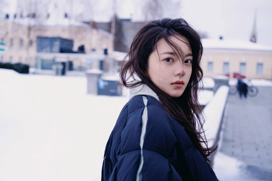 赵今麦身着藏蓝羽绒服，在积雪覆盖的户外回头望向镜头，背景是虚化的欧式建筑。