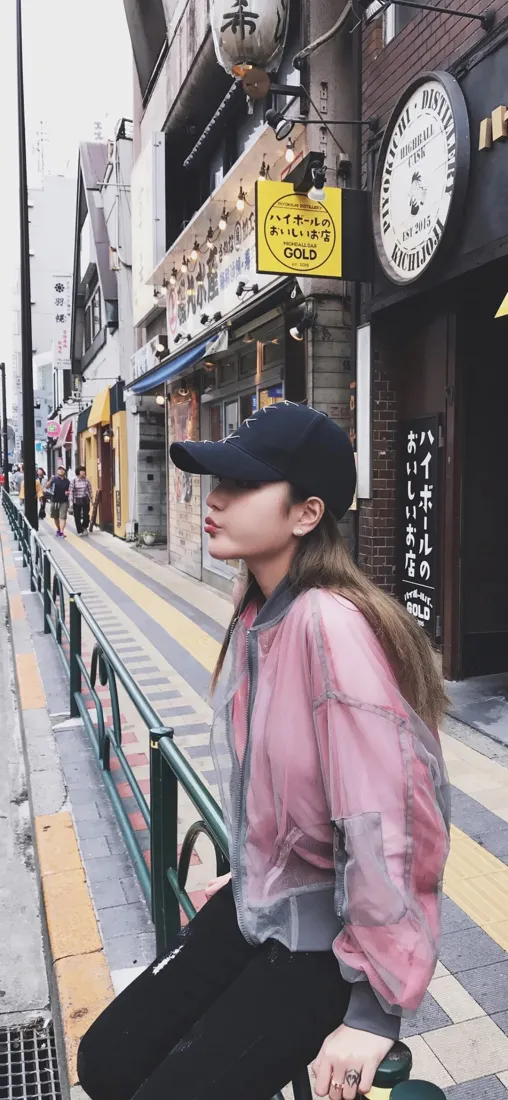 戴黑棒球帽、穿粉灰拼色外套的女生侧身站在日式商业街人行道上