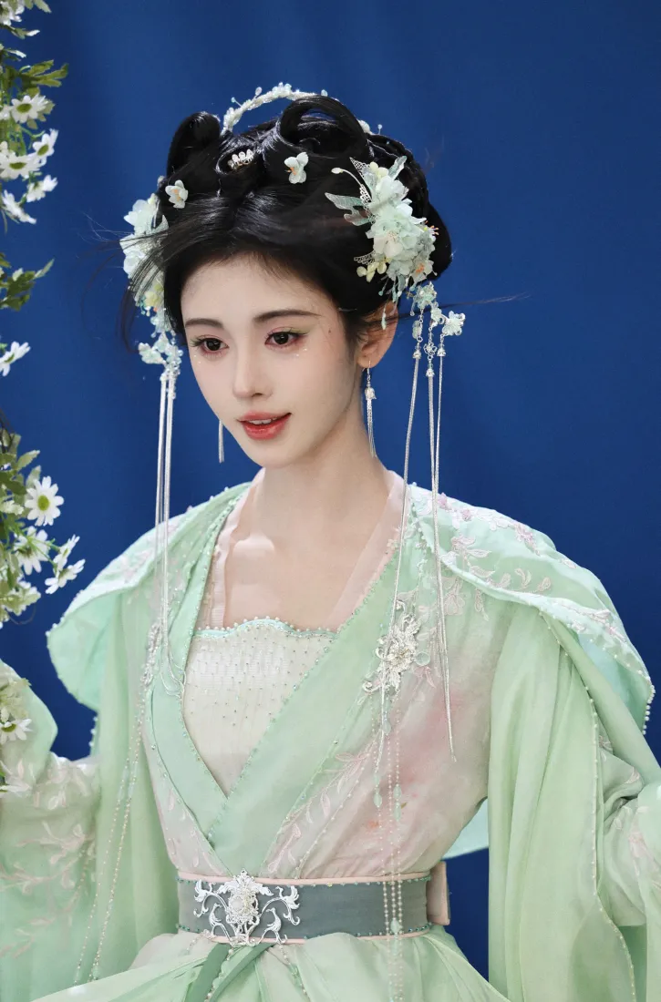 鞠婧祎着浅绿纱质古装，头戴白花流苏发饰，立于纯蓝背景前，身侧有白色小花枝。