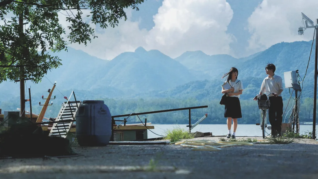 湖畔码头边，穿JK制服的女生与扶自行车的男生并肩，远处是层叠青山与蓝天白云。
