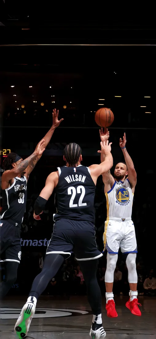 NBA赛场中库里身着勇士队服跃起投篮，两名防守球员抬手封堵。