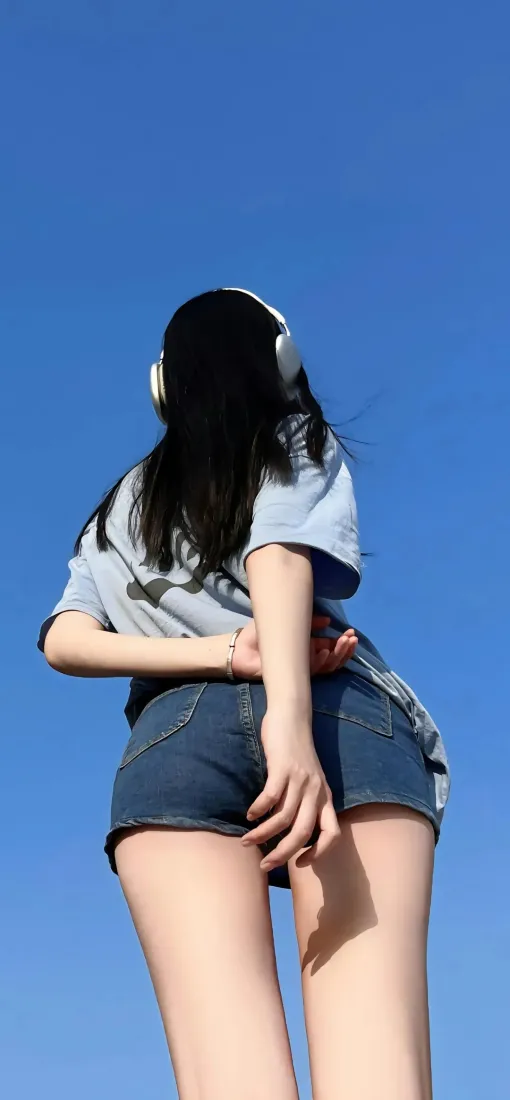 低角度仰拍戴白色耳机的长发少女背影，身着灰T恤与牛仔短裤，背景为纯净蓝天