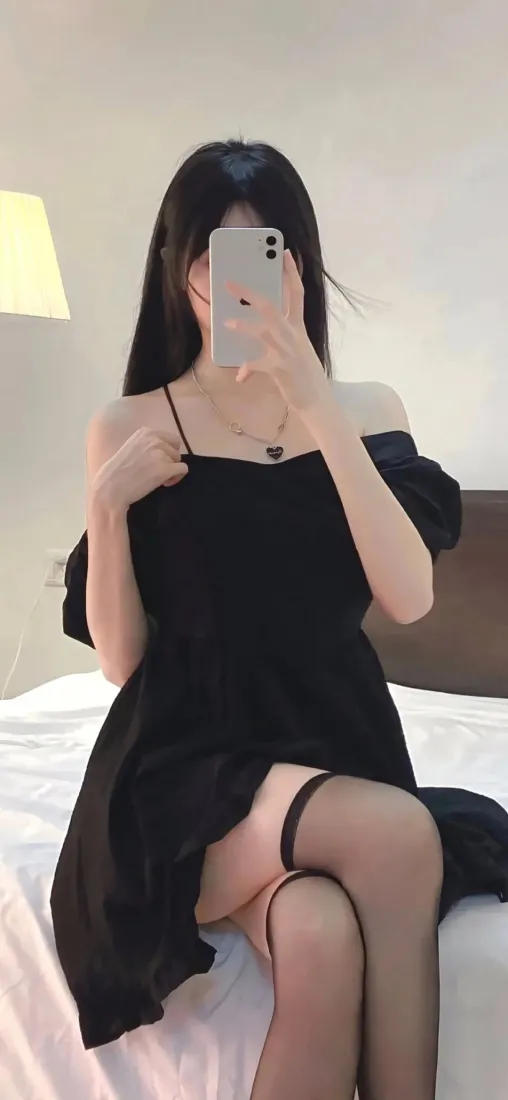 长发女子穿黑色露肩吊带裙与黑丝，坐床上用手机对镜自拍。