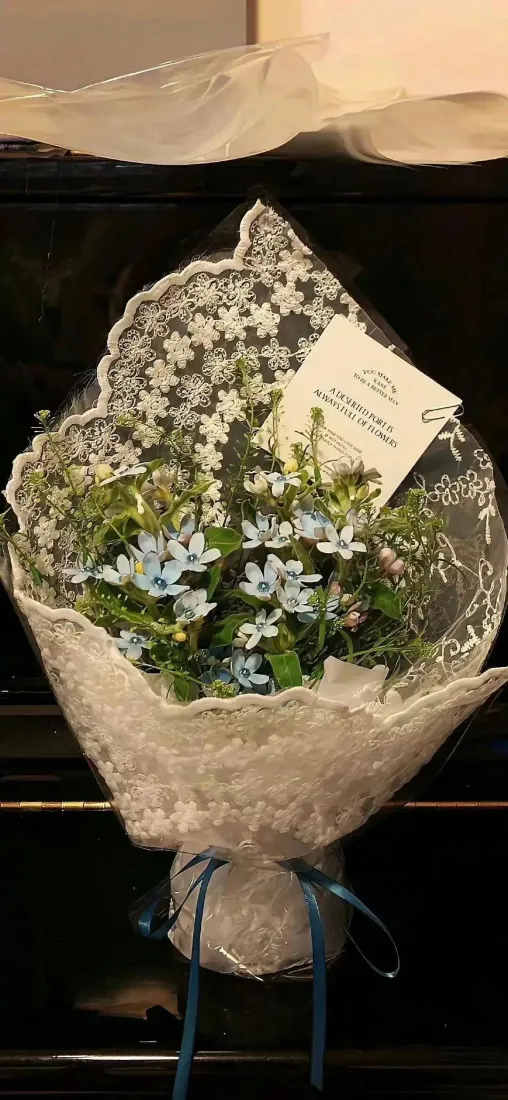 白色蕾丝包装的蓝绿色小花束，系蓝色丝带，附白色卡片，置于深色背景前