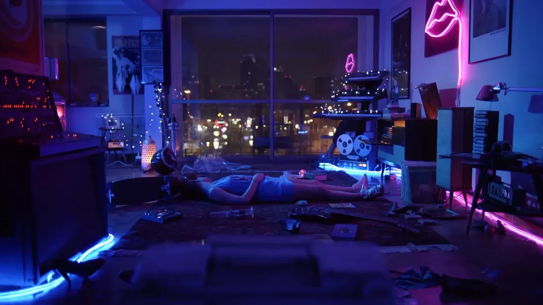 霓虹灯光点缀的夜晚房间，一人躺于地板，窗外是城市夜景