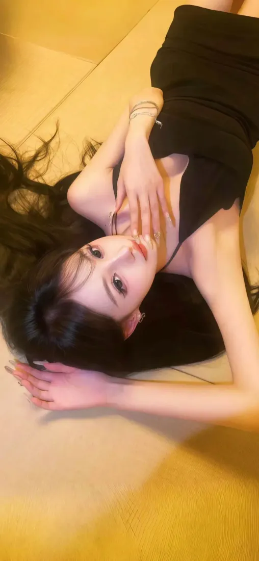 长发年轻女子身着黑色抹胸裙，躺于暖黄色调室内地面，姿态柔媚。