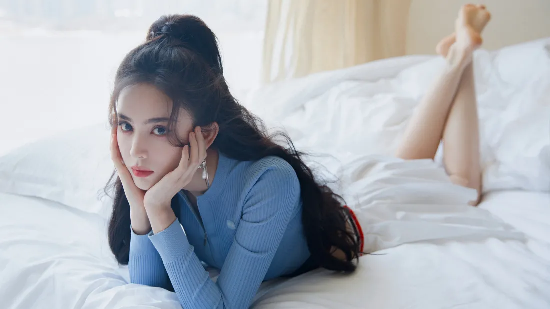 长发女子身着蓝色上衣，双手托腮趴卧在铺有白色床品的床上。