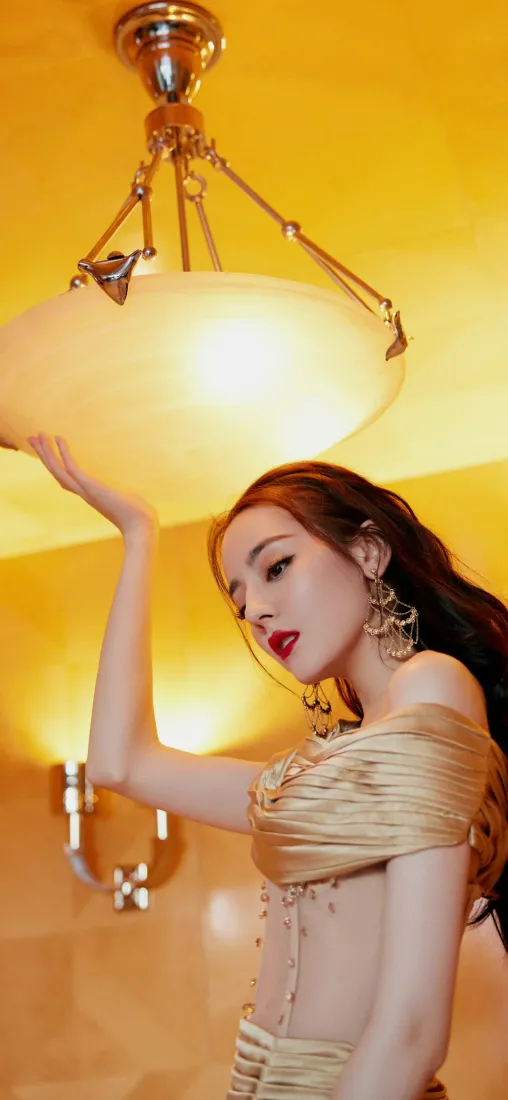 暖黄灯光下迪丽热巴身着金色露肩礼服，抬手轻托复古吊灯灯罩，造型优雅。