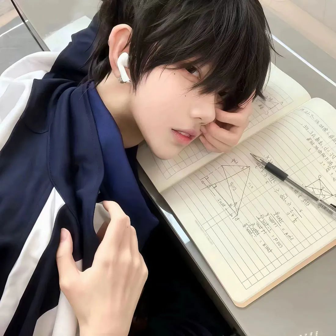 穿蓝白校服的短发男生趴在课桌笔记本旁，戴白色无线耳机，侧脸看向镜头。