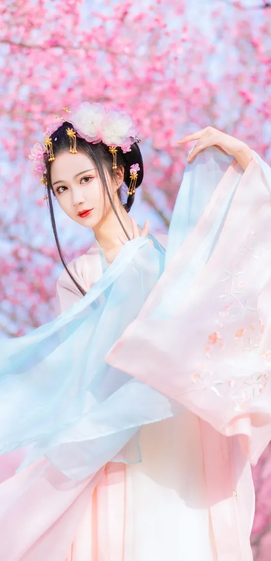 身着粉蓝渐变汉服的女子立于盛放的粉色樱花树下，抬手轻扬衣袖。