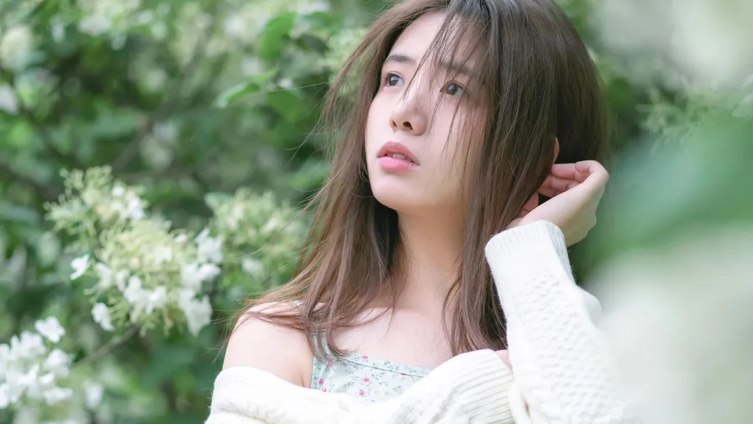 长发年轻女子身着白色针织衫，置身绿意花丛中抬手轻触耳侧发丝。