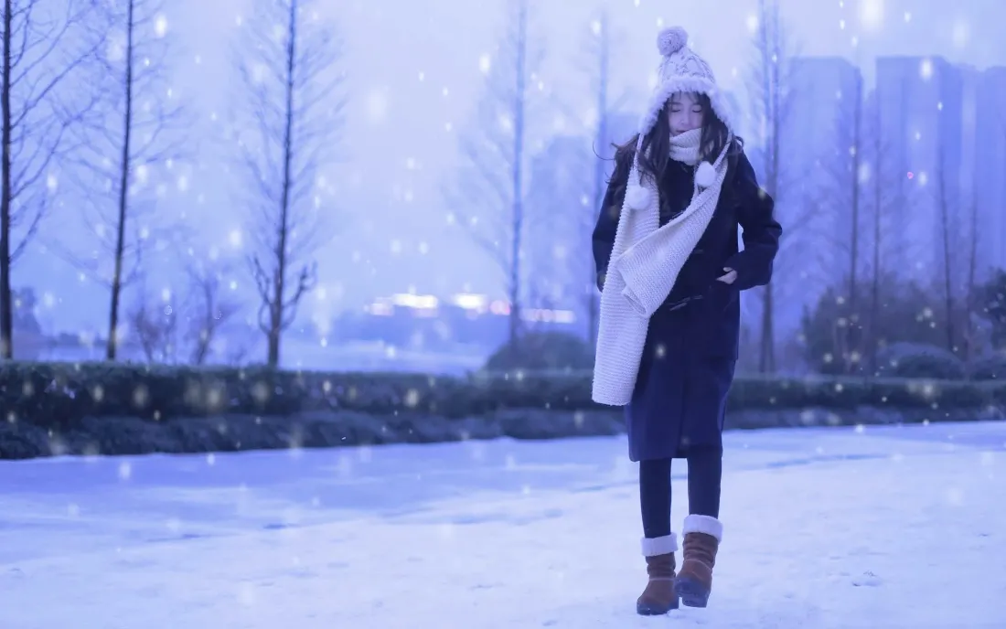 飘雪冬日户外，戴白帽围长围巾的黑衣女子站在积雪地面上。