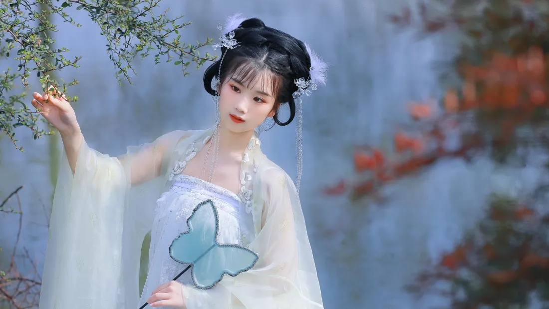 身着浅白汉服的女子手持蝴蝶团扇，抬手轻触枝桠，背景朦胧虚化。