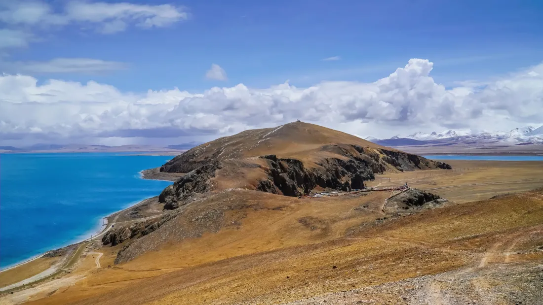 西藏高原圣湖风光，湛蓝湖水环绕褐黄色山丘，远处雪山与白云相映。