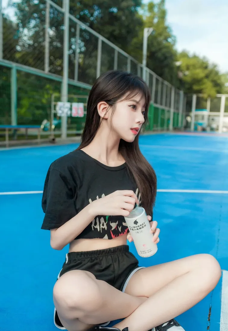 长发黑衣少女盘腿坐在蓝色球场，手持带吸管的白色饮品杯侧头望向画面右侧