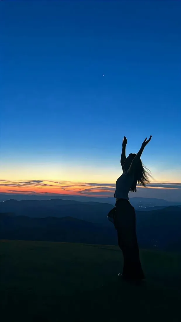 山顶黄昏时分，女性剪影立于画面右侧，背景是渐变蓝橙天空与层叠远山。