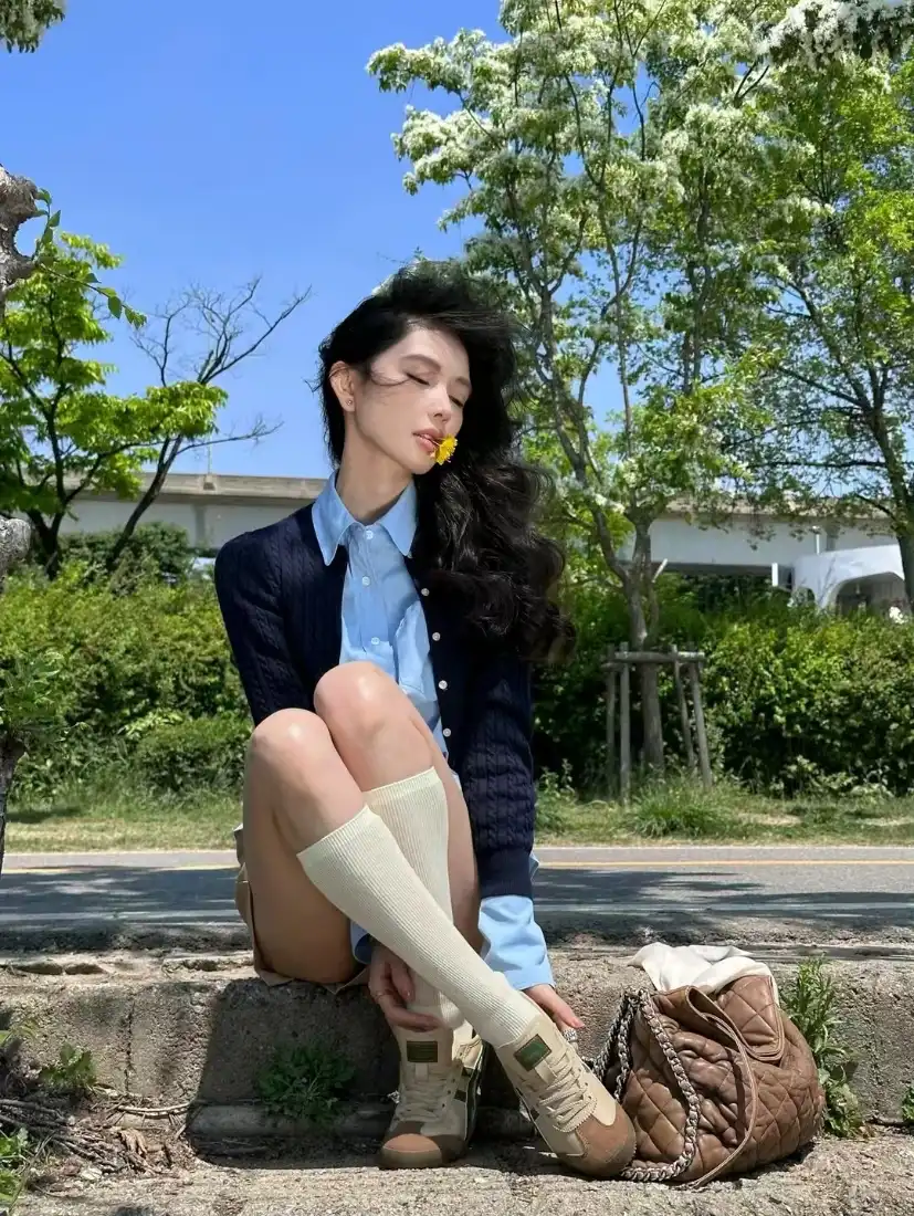 卷发女子坐路边石阶，嘴叼黄花，蓝衬衫外搭深色开衫，背景绿树蓝天。