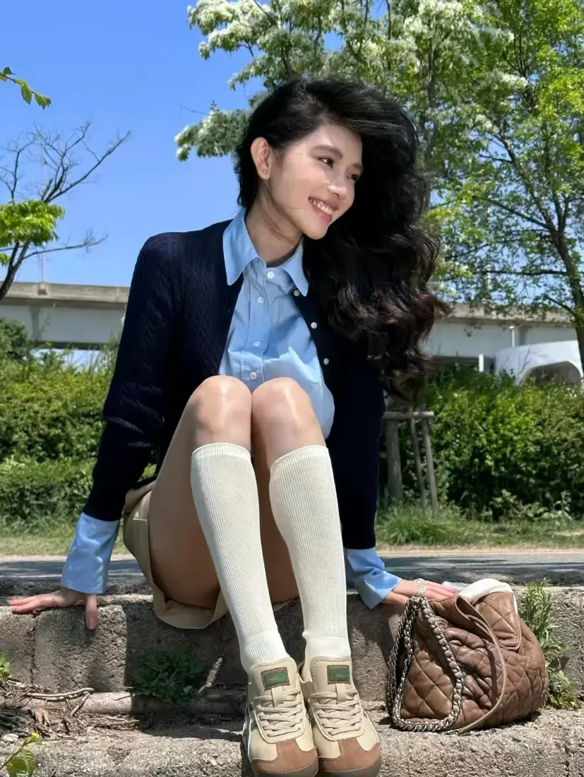 长发青春女孩坐在户外石阶上，穿休闲学院风穿搭，背景是蓝天绿树。