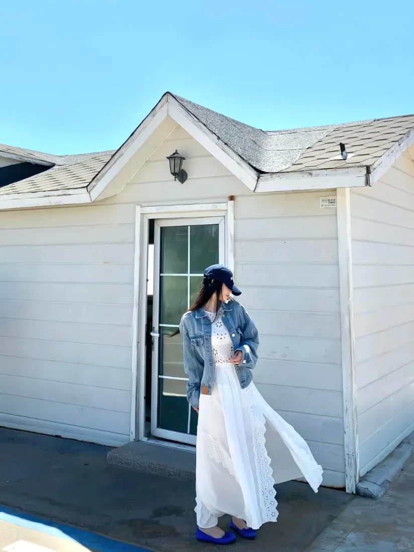 白衣牛仔外套女子站在白色小屋前，蓝天背景清新明亮。