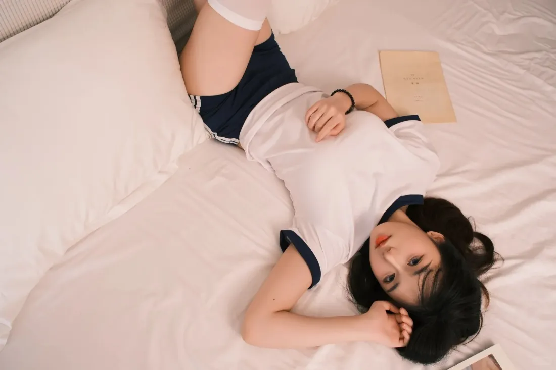 2k卧室真人壁纸|居家背景图片 - 安逸「哲风壁纸」 短发年轻女子身着白短袖蓝短裤,躺于铺白色床品的卧室床上。