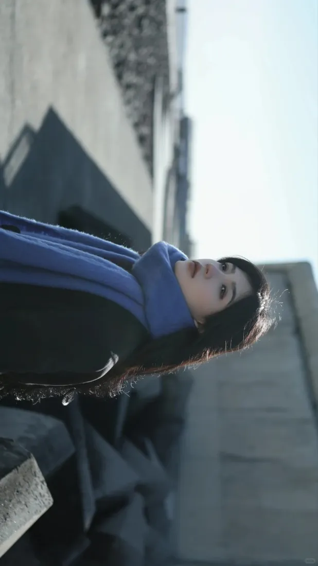 户外低角度拍摄，戴蓝色围巾的长发女性，背景为虚化的现代建筑与明亮天空。