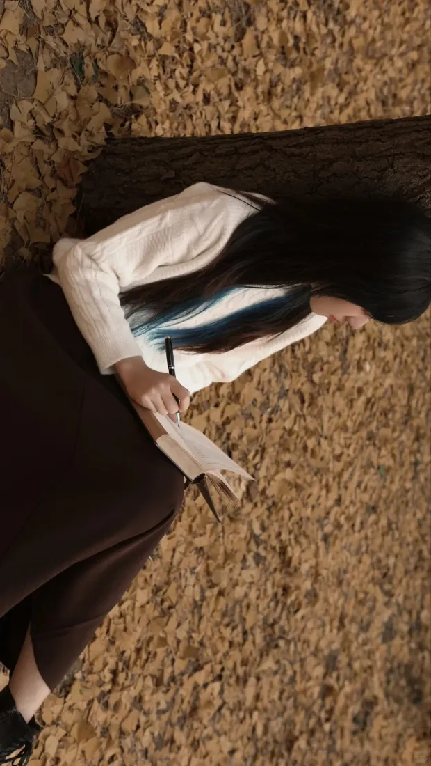 俯拍秋日铺满落叶的地面上，长发女生坐于树干旁持纸笔书写