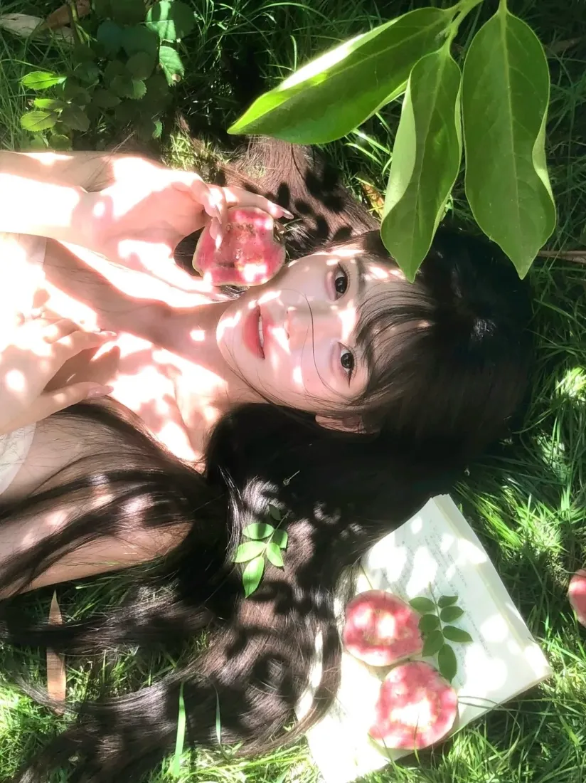 戴眼镜的长发女生躺于草地，手持粉花，树影光斑洒落，氛围清新浪漫