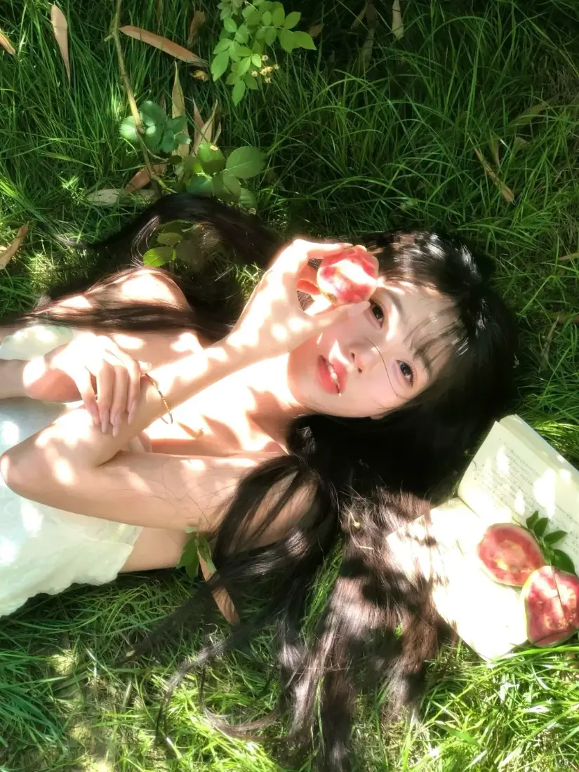 长发白衣女子躺于青草地，手持红玫瑰，树影光斑洒落，氛围清新浪漫。