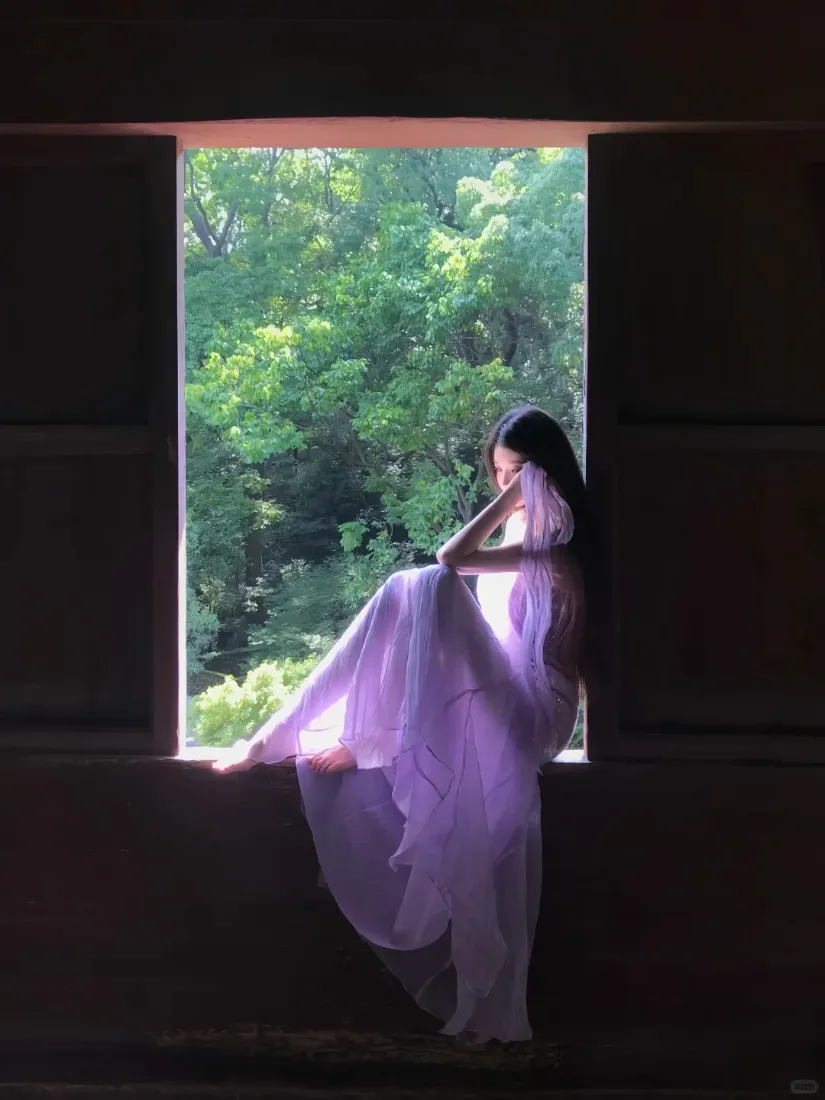 身着淡紫色纱裙的女子坐于窗边，窗外是葱郁的绿树，室内光线昏暗。