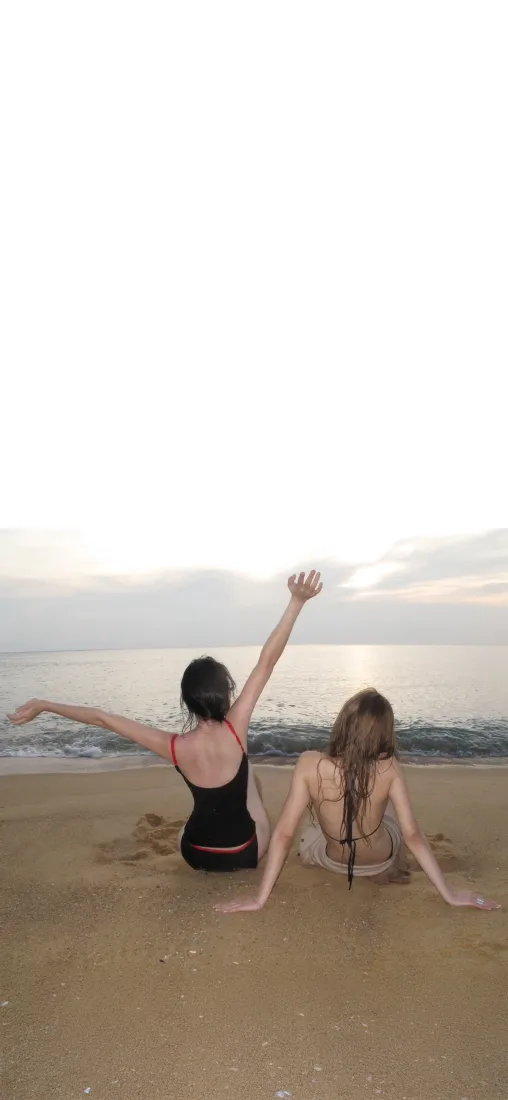 两名身着泳装的女子背对镜头坐在黄昏沙滩上，面朝海面，一人手臂舒展抬起。