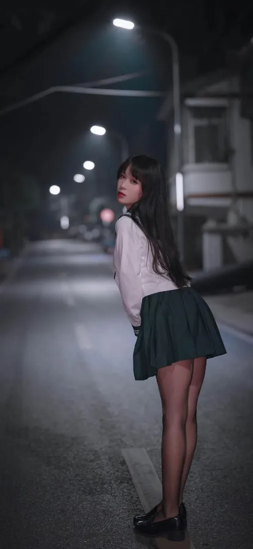 夜晚街道上，穿白衬衫绿短裙黑丝的长发少女侧身回头望向镜头，路灯沿街道向远处延伸。