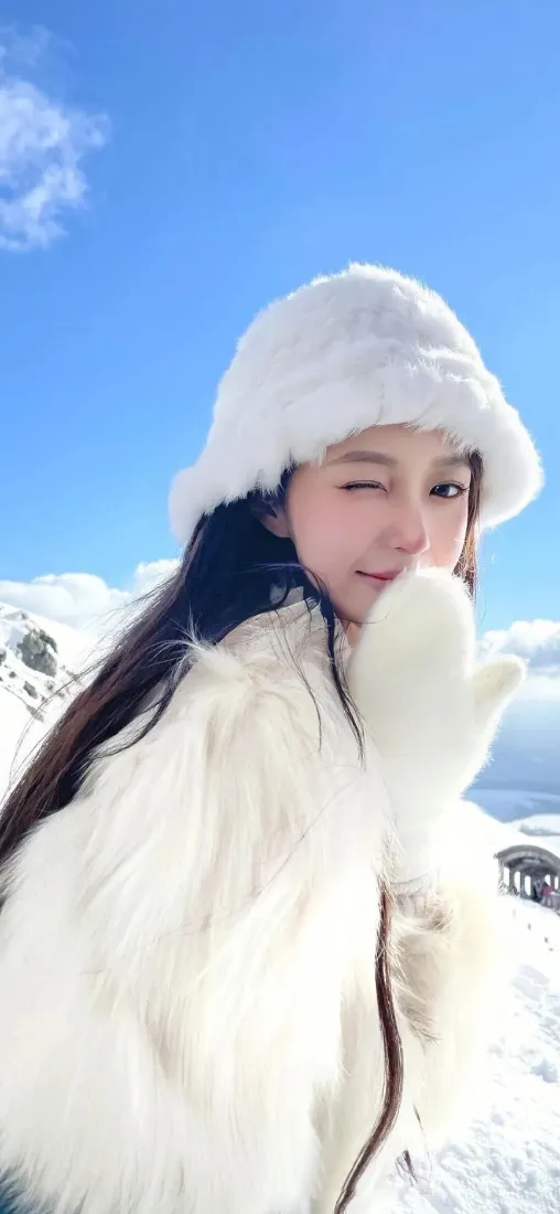 身着白色毛绒服饰的长发女生在雪山蓝天背景前俏皮眨眼自拍