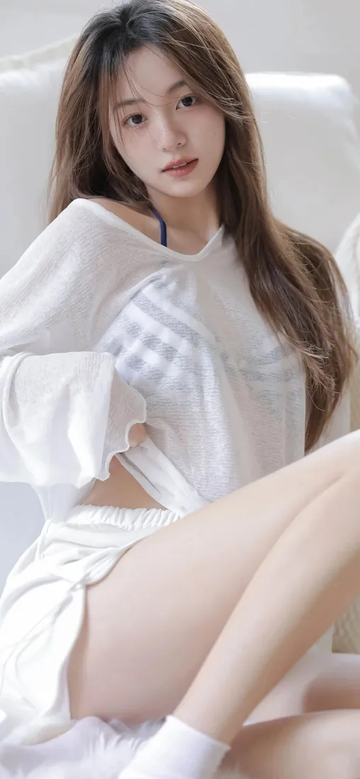 长发清纯女生身着宽松白衣居家坐姿，背景为浅色沙发，氛围柔和清新。