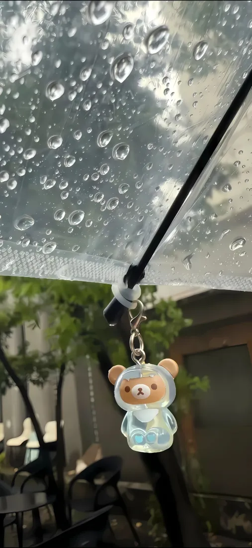 雨天透明伞面挂着小熊挂件，背景虚化呈现绿植与建筑。