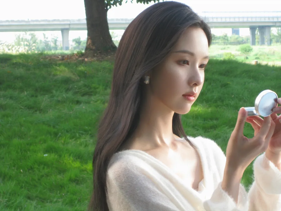 白衣长发女子在户外草地手持圆形化妆镜补妆，背景虚化有树木与高架桥