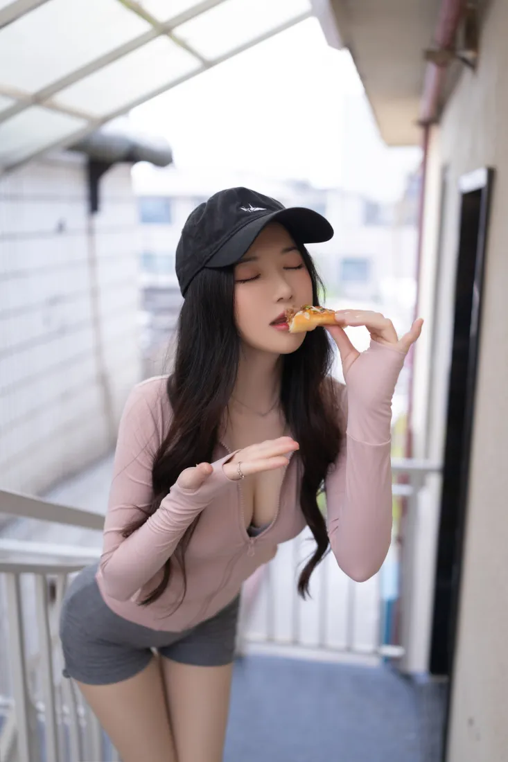 戴黑色棒球帽的长发女生在户外阳台，穿粉色上衣灰色短裤，正吃手中的食物。