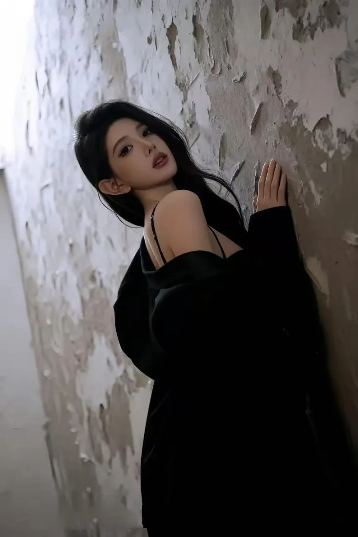 长发黑衣美女侧身靠在斑驳剥落的墙前，回头看向镜头。