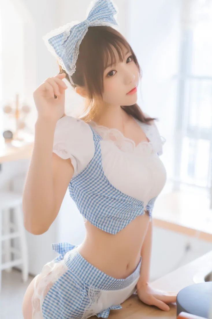 明亮厨房中，穿蓝白格纹露腰厨娘装、戴同色系大蝴蝶结的年轻女生侧身倚靠台面。