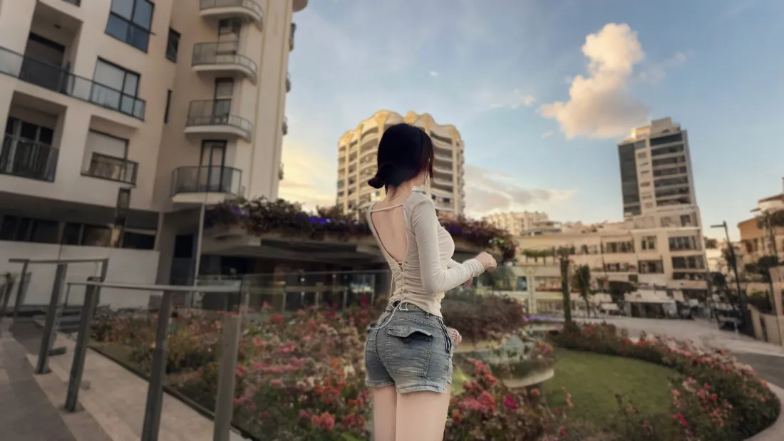 白衣露背短发女子背对镜头立于城市花园，背景为高层住宅与黄昏天空。