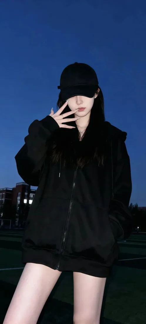 戴黑帽穿黑色连帽外套的女生，在深蓝色夜空下抬手比出手势。
