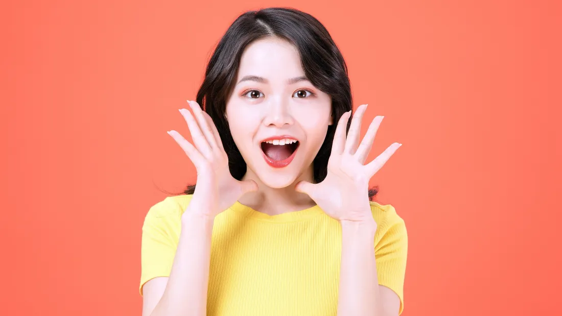 4k黄衣女生壁纸|红色背景 - 可爱迷人「哲风壁纸」 橙红纯色背景前,黄衣黑发女生双手举在脸颊旁,张嘴露惊喜神态。
